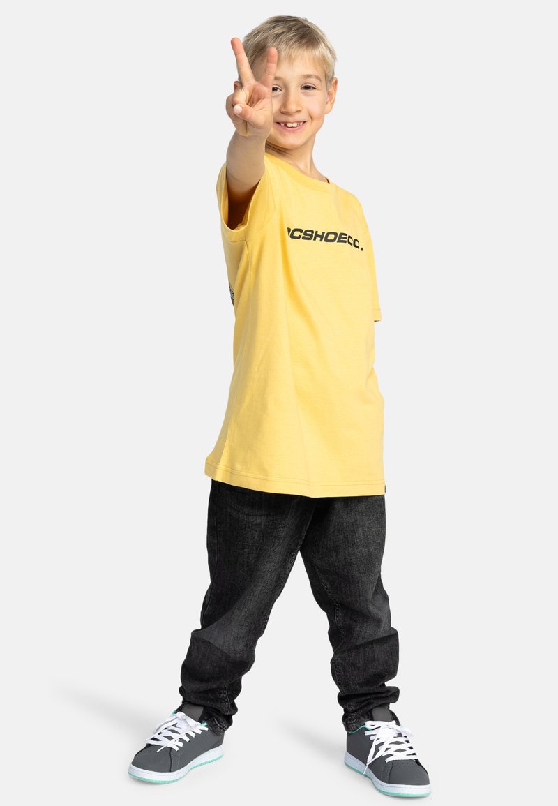 Camiseta de algodón amarilla con el logo "DCSHOECO.", combinada con pantalones de mezclilla oscuros y zapatillas grises con cordones blancos y suelas azul claro.
