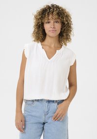 Haut sans manches blanc avec un décolleté en V, des bordures froncées et un léger plissé. Associé à un jean en denim taille haute bleu clair.