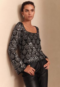 Čierny top s flitrami so štvorcovým výstrihom, zdobený vzorom v tvare mušle, s dlhými rukávmi, doplnený čiernymi koženými nohavicami.
