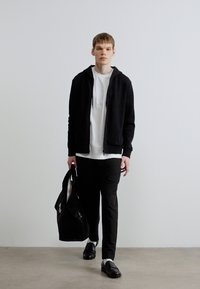 Črna jopa z zadrgo preko bele majice, črne hlače in črni mokasini. Model nosi črno torbo z belimi poudarki.