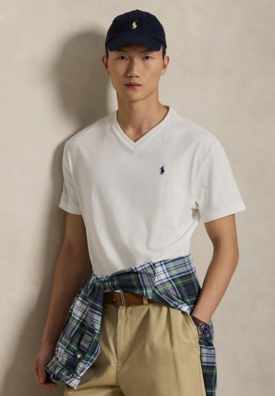 White cotton v-neck t-shirt with navy polo logo, beige pants, and a plaid shirt tied around the waist. Navy cap with polo logo.