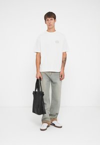 Witte katoenen t-shirt met een rond embleem op de borst, gecombineerd met lichtgewassen denim jeans en witte sneakers. Houdt een zwarte tote bag vast.