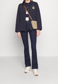 Marinblå vadderad jacka med knappstängning framtill och ett emblem, bärs över en randig skjorta och kombineras med mörka utställda jeans och cremefärgade sneakers.