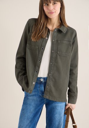 Kobieta ubrana w oliwkowozieloną kurtkę na guziki, na białej bluzce i niebieskich dżinsach, trzymająca pasek brązowej torebki jedną ręką.