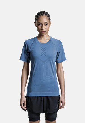 Femme aux cheveux tressés portant un t-shirt de sport bleu à manches courtes et un short de sport noir superposé, debout face à l'avant sur un fond blanc.