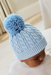 Lichtblauwe gebreide muts met kabelpatroon en een pluizige pompon bovenop. Geribbelde band aan de onderkant voor een stevige pasvorm.