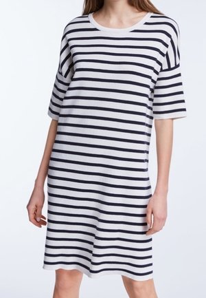 Vrouw die een witte knielange jurk draagt met zwarte horizontale strepen en ellebooglange mouwen, tegen een effen achtergrond.