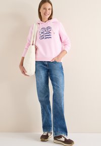 Hoodie rose avec un graphisme "sea", jean évasé en denim bleu, et chaussures de sport marron. Accessoires : un sac épaule matelassé blanc.