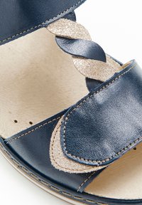 Sandalia de cuero en azul marino con un acento trenzado metálico dorado. Presenta detalles de costura y un diseño abierto. Textura suave, estilo casual.