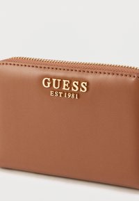 Portofel maro din piele eco cu logo auriu „GUESS EST 1981”, dispune de închidere cu fermoar și suprafață texturată, detalii cu cusături contrastante.