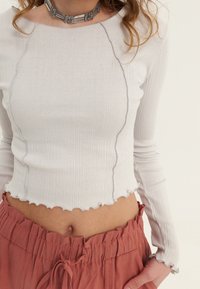 Mujer que lleva una camiseta de manga larga de canalé en color crema y pantalones de tiro alto en color óxido con cintura ajustable, que muestran parte de su abdomen.