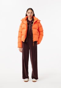 Giacca a vento arancione con colletto alto, abbinata a un completo in velluto marrone scuro con piping bianco e zip. Sneakers bianche completano il look.