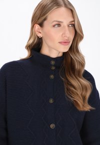 Granatowy, dziergany kardigan z wysokim kołnierzem, teksturowanym wzorem w zygzaki oraz pięcioma brązowymi guzikami z przodu. Miękki i przytulny materiał.