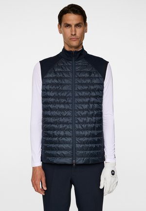 Navy-blau gesteppte Weste mit hohem Kragen, Reißverschluss und strukturierter Oberfläche, kombiniert mit einem weißen Langarmshirt und einem Handschuh.