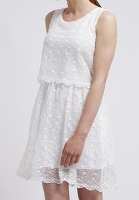 Weißes Spitzenkleid mit einem enganliegenden Oberteil, das einen runden Ausschnitt und florale Stickereien aufweist, sowie einem ausgestellten Rock. Der Spitzenabschluss fügt Textur und Detail hinzu.