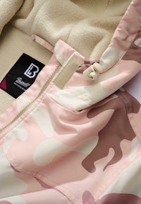 Rožinės ir smėlio spalvos kamufliažinė striukė su vilnos pamušalu, su užtrauktuku, lipnia juosta užsegimu ir juodai balta prekės ženklo etikete.