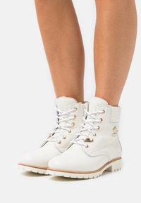 Bottes de cheville en cuir blanc avec œillets en métal, bouts ronds et semelle beige contrastante. Présente une marque dorée sur le côté.