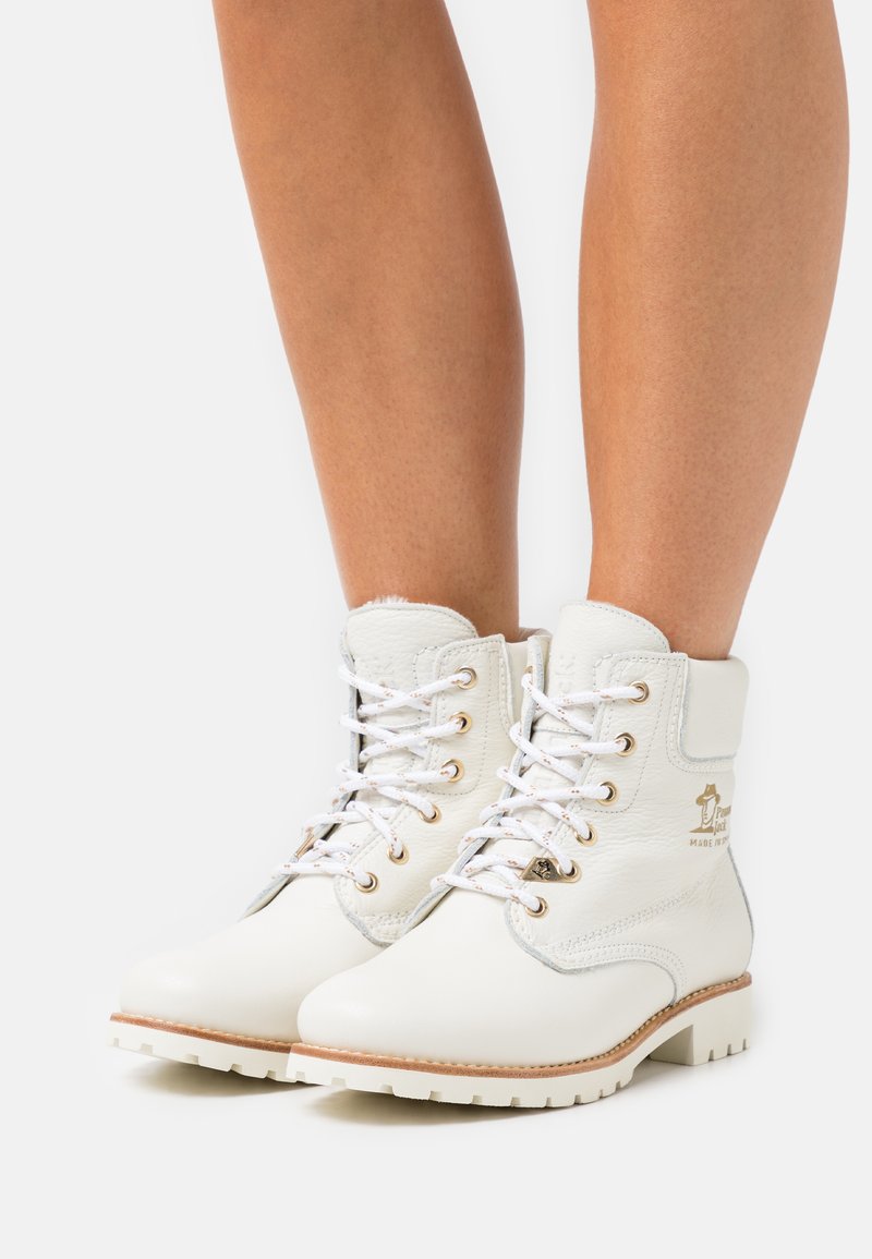 Bottes de cheville en cuir blanc avec œillets en métal, bouts ronds et semelle beige contrastante. Présente une marque dorée sur le côté.