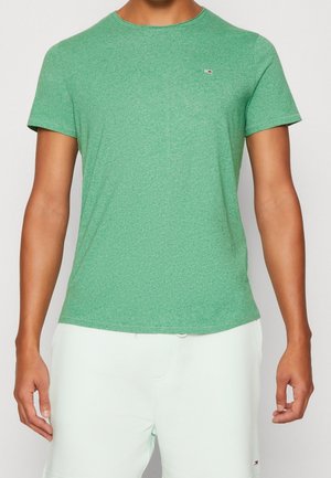 Uomo che indossa una maglietta verde attillata a maniche corte e pantaloni di colore chiaro, con un piccolo logo sul petto e sui pantaloni.