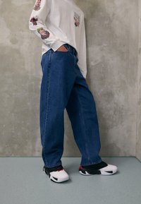 Jeans de mezclilla azul de pierna ancha, camiseta de manga larga blanca con gráficos coloridos en las mangas, y zapatillas deportivas en blanco y negro. Fondo de hormigón.
