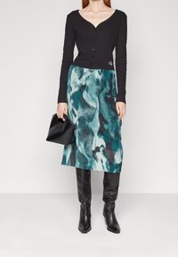 Svart långärmad knäpptröja, teal mönstrad midikjol med nyanser av svart, knähöga svarta läderstövlar och en liten svart handväska.