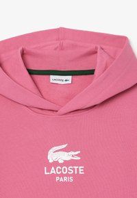 Felpa rosa con cappuccio regolabile, logo e testo bianchi Lacoste, dettaglio verde all'interno del colletto, realizzata in morbido materiale di cotone.