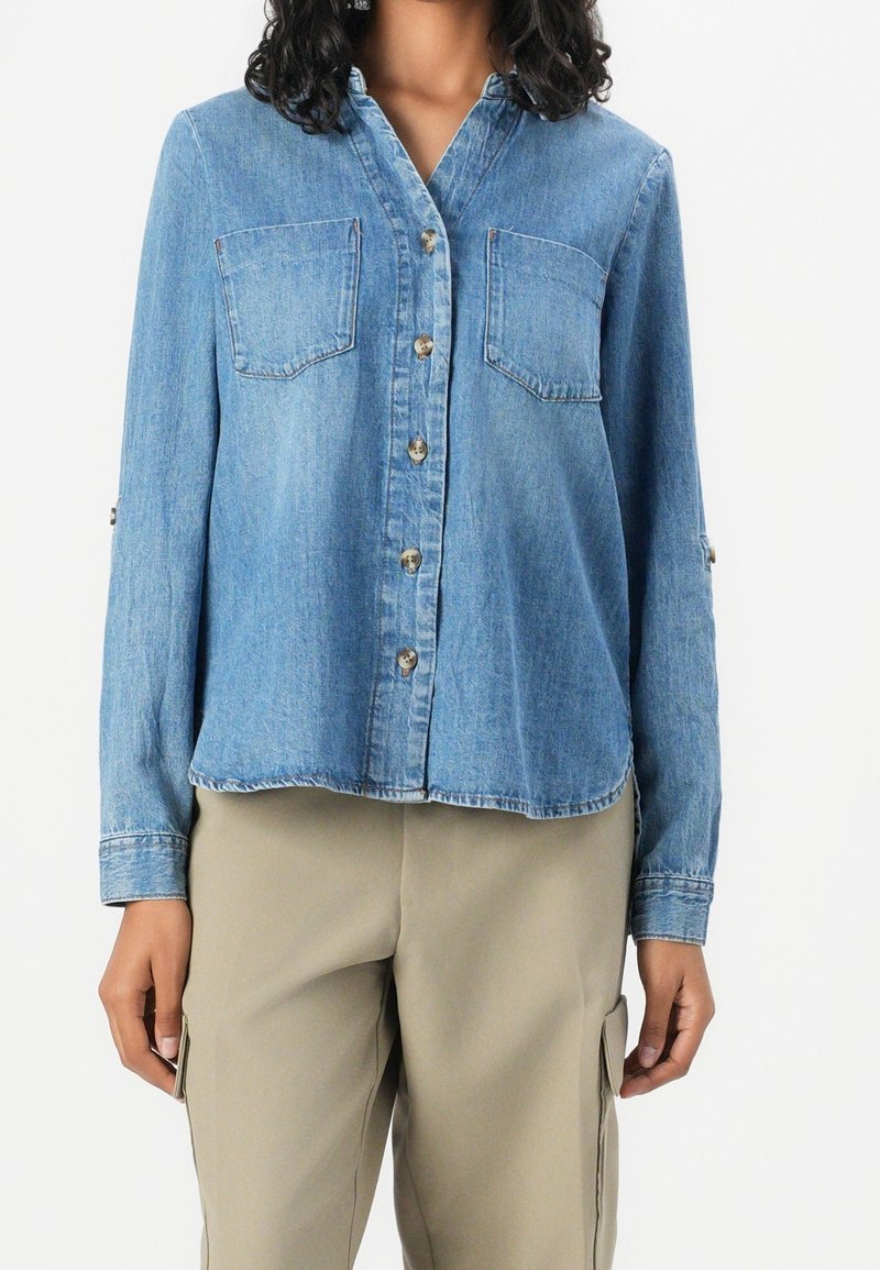 Femme portant une chemise en denim bleu à manches longues boutonnée avec deux poches poitrine et un pantalon beige avec des poches latérales.