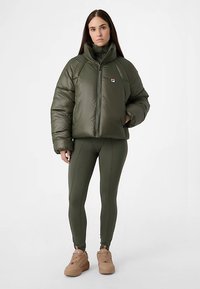 Giacca a piumino verde con collo alto e chiusura con zip, abbinata a leggings coordinati e sneakers color cammello. Il tessuto è liscio e imbottito.