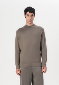 Maglione lungo marrone a maniche lunghe con scollo alto e texture a costine. Indossato con pantaloni grigi abbinati, l'outfit presenta un look coeso e minimalista.