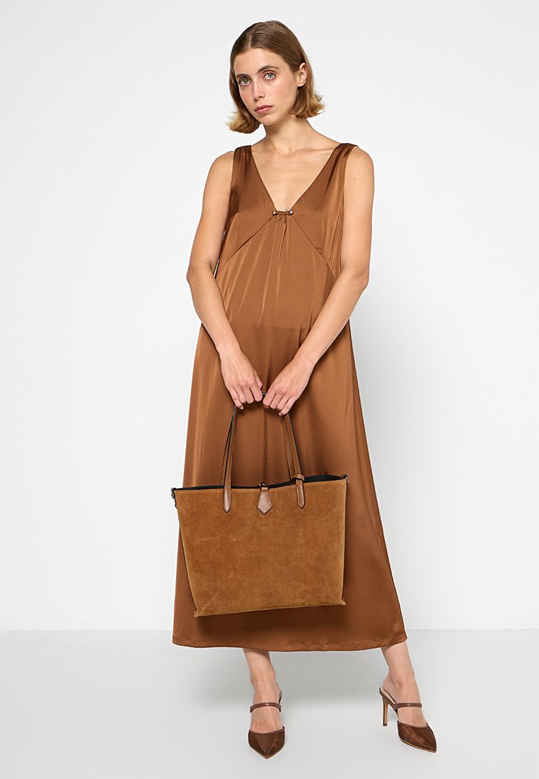 Braune Wildleder-Tasche mit strukturiertem Finish, schwarer Innenausstattung und doppelten Griffen. Das Model trägt ein passendes braunes ärmelloses Kleid.