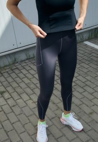 Czarne legginsy z kontrastującymi szarymi szwami i siateczkowymi akcentami. W połączeniu z białymi butami sportowymi z neonowo zielonymi detalami.