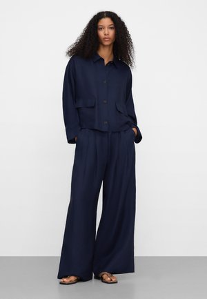 Frau mit lockigem Haar, die ein marineblaues Hemd mit Knöpfen und weit geschnittene Hose trägt, steht mit den Händen in den Taschen und trägt schwarze Sandalen.