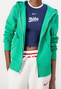 Grön zip-up hoodie med snoddar, tillverkad av mjukt tyg. Bärs över en marinblå crop top med vit Nike-logotyp, i kombination med gr å sweats.