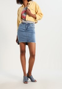 Nainen, jolla on päällään keltainen lyhyt takki, harmaa t-paita, jossa on punainen huulipainatus, farkku mini-hame ja siniset avonaiset korkokengät, seisoo.