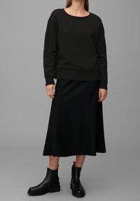Schwarzer Strickpullover, langer schwarzer Rock und schwarze Knöchelschuhe. Der Pullover hat einen runden Halsausschnitt und eine lässige Passform. Insgesamt glatte Textur.