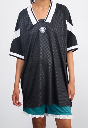Personne portant un maillot de sport Nike noir et blanc oversized par-dessus un short bleu sarcelle avec une bordure en dentelle blanche sur un fond uni.