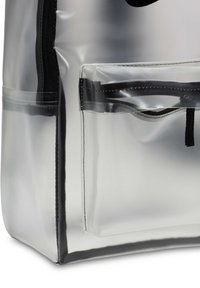 Gjennomsiktig PVC totebag med en blank overflate, som har en frontlomme og svarte glidelåser. Kantene er sydd med svart tråd.