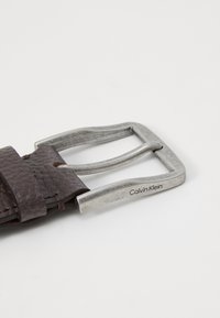 Ceinture en cuir brun foncé avec une surface texturée et une boucle en métal argenté. La boucle présente le logo "Calvin Klein".