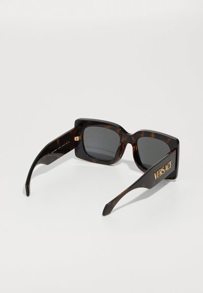 Tortijas saulesbrilles ar drosmīgu kvadrātveida formu, tumši pelēkiem lēcu un zelta "Versace" zīmolu uz tempļiem.