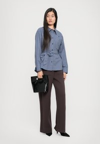 Sinine ruuduline särk, mille vöökoht on kinni seotud, koos tumedate pruunide laiadest pükstest. Käes must läikiv käsipagas.