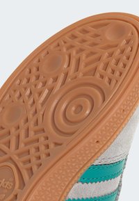 Sneakerzool met een gestructureerd lichtbruin rubberen oppervlak, voorzien van circulaire en geometrische patronen, aangevuld met grijs suede en teal strepen.