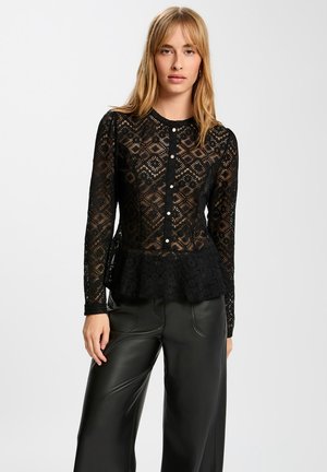 Femme blonde portant un haut péplum à boutons en dentelle noire et un pantalon en cuir noir, debout devant un fond blanc uni.