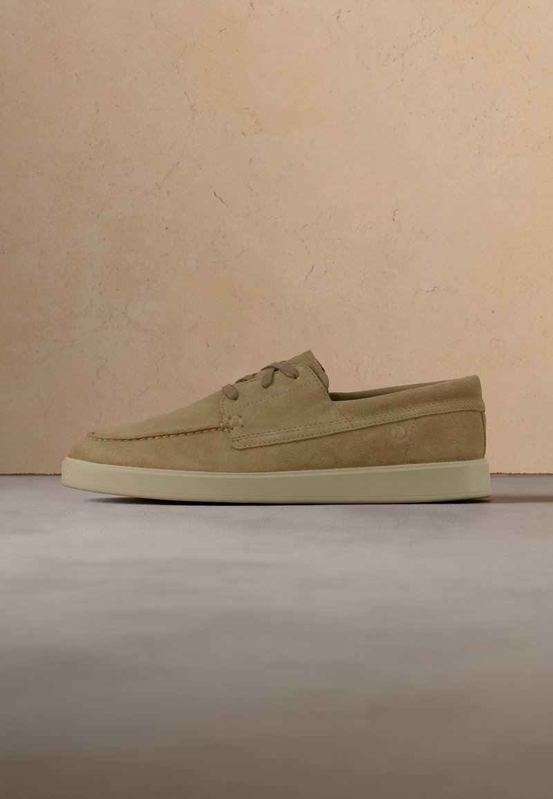 Chaussure décontractée en daim marron clair avec lacets et semelle en caoutchouc beige sur une surface grise devant un mur texturé beige.
