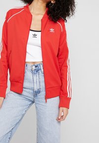 Červená zipová bunda s bílými pruhy na rukávech, nošená přes černý a bílý crop top, spárovaná s světle modrými džínami s vysokým pasem.