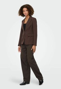 Blazer marrontailleur avec deux poches avant, porté sur un haut noir. Associé à un pantalon large marron foncé et des bottes à talons noires.