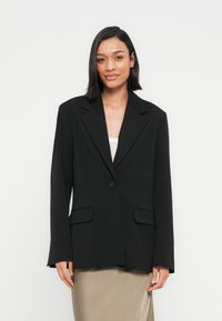 Γυναίκα με μαύρα μαλλιά μέχρι τους ώμους, που φοράει ένα υπερμεγέθες μαύρο σακάκι πάνω από λευκό τοπ και μπεζ φούστα, μπροστά σε απλό ανοιχτό φόντο.