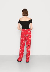 Top preto off-the-shoulder com bordas com franzido e calças com padrão floral vermelho; modelo de costas, mostrando um corte relaxado e uma textura suave.