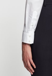 Mano descansando al lado, vistiendo una camisa blanca de botones y pantalones negros texturizados contra un fondo claro y liso.