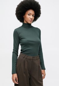 Frau mit natürlicher Afrofrisur, die einen dunkelgrünen Rollkragenpullover und braune, hoch taillierte, plissierte Hosen trägt und vor einem schlichten weißen Hintergrund steht.