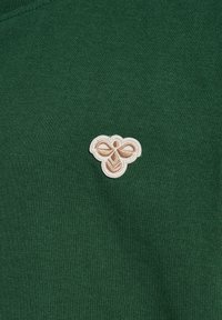 Grön skjorta med en krämfärgad broderad bi-design på vänster bröst. Tyget har en slät, strukturerad yta.